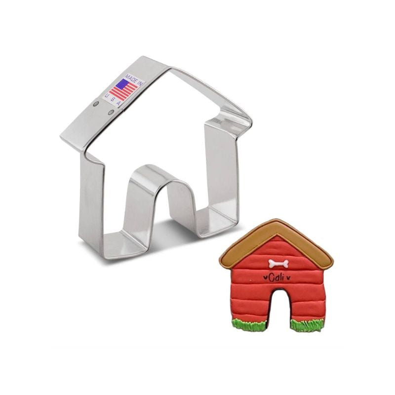 A metal cookie cutter in the shape of a house, labeled "Made in U.S.A." with a small American flag, is next to a decorated dog house-shaped cookie.