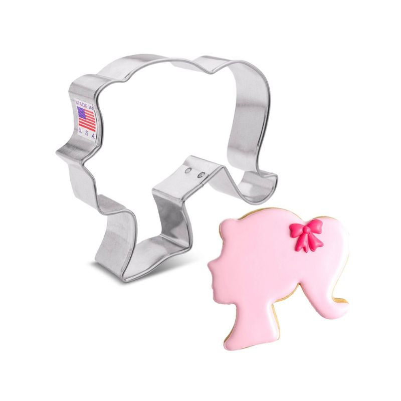 Metal cookie cutter shaped like a doll head beside a pink sugar cookie with pink bow