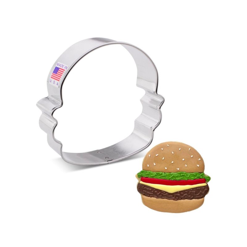 Metal cookie cutter made in the usa with American flag sticker next to a decorated cookie hamburger with bun and green lettuce icing tomato and icing burger patty