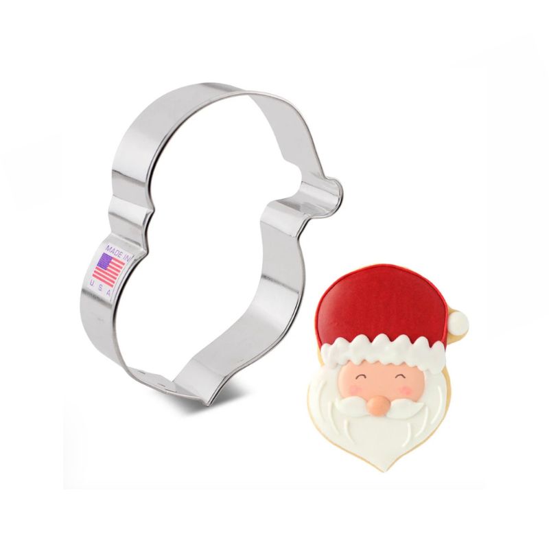 Stainless steel Santa face cookie cutter with American flag sticker beside sugar cookie santa face with red hat and white beard and cute eyes