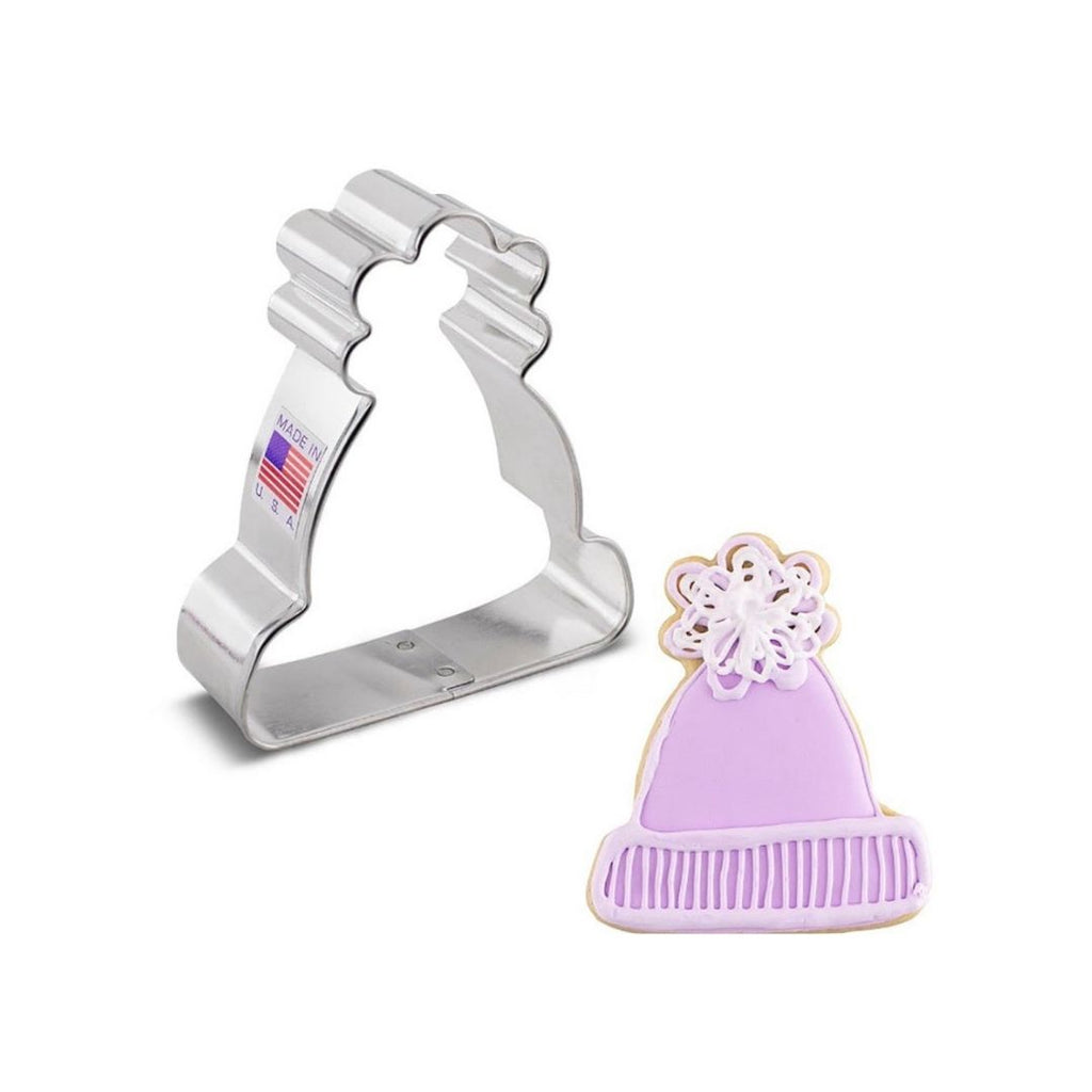 Metal cookie cutter in the shape of a winter hat that has a made in the usa with flag sticker on side next to a sugar cookie with purple royal icing on top