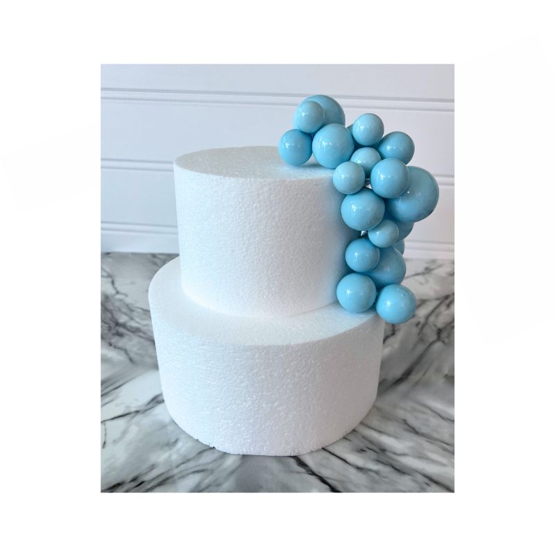 A two-tier white cake showcases a cascading cluster of Cake Sphere multi-sized balls in Baby Blue on one side. The cake is set against a backdrop with a white wainscot panel and rests on a marble surface.