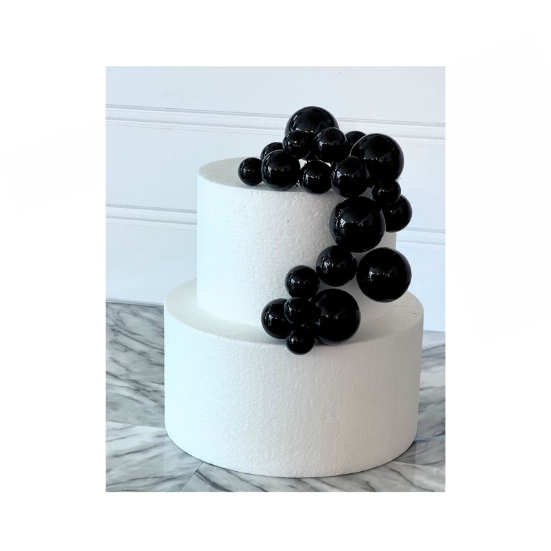 A two-tier white cake showcases a cascading cluster of Cake Sphere multi-sized balls in shiny Black on one side. The cake is set against a backdrop with a white wainscot panel and rests on a marble surface.