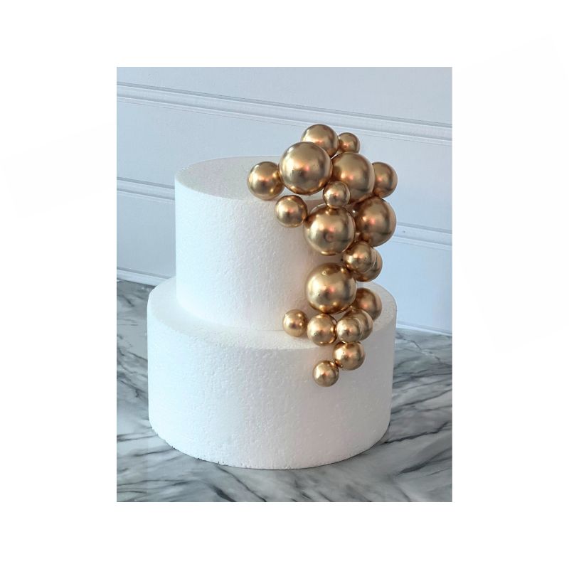 A two-tier white cake showcases a cascading cluster of Cake Sphere multi-sized balls in shiny gold on one side. The cake is set against a backdrop with a white wainscot panel and rests on a marble surface.