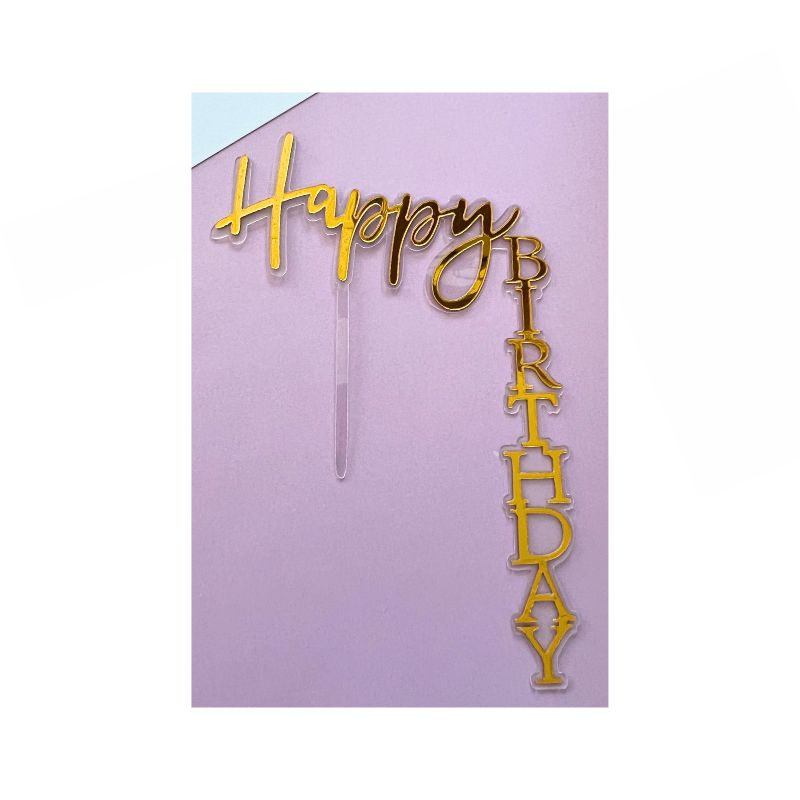 The Gold Happy Birthday Cake Topper features "Happy" in cursive script positioned horizontally and "BIRTHDAY" in block letters vertically, all set against a light pink background. The topper is designed with a clear stick to be easily placed into the cake.