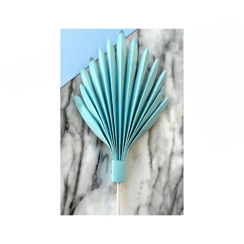 A Cake Paper Palm, fully opened with neat, symmetrical accordion folds, rests on a marble surface. The light blue, folded paper fan with a handle partially overlaps a corner of pastel blue paper.