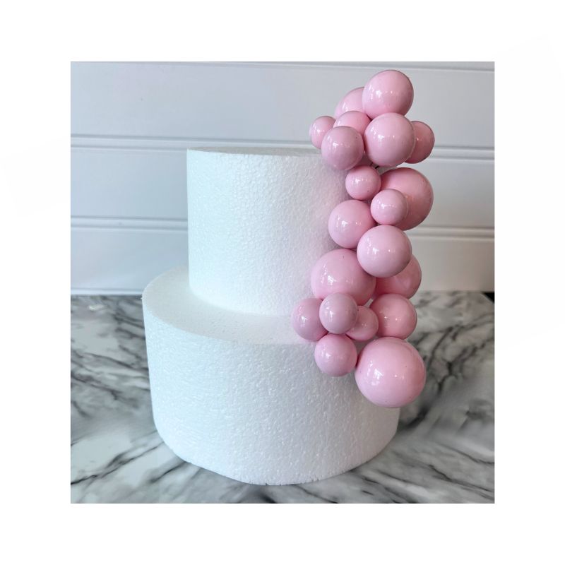 A two-tier white cake showcases a cascading cluster of Cake Sphere multi-sized balls in baby pink on one side. The cake is set against a backdrop with a white wainscot panel and rests on a marble surface.