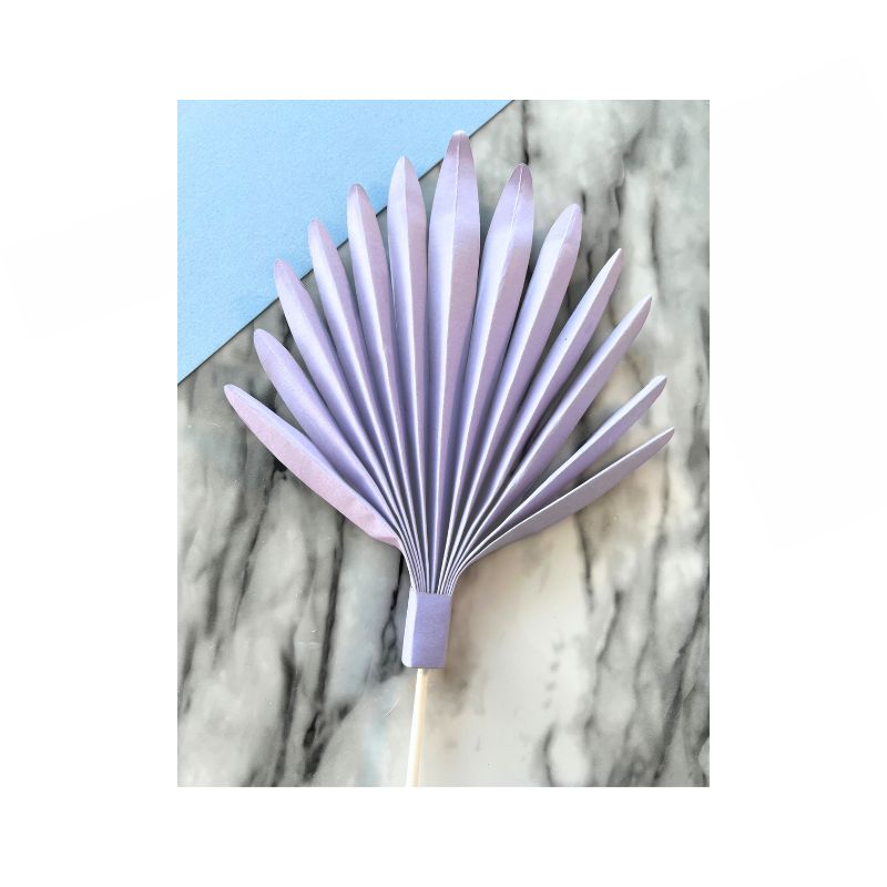 A Cake Paper Palm, fully opened with neat, symmetrical accordion folds, rests on a marble surface. The light purple, folded paper fan with a handle partially overlaps a corner of pastel blue paper.