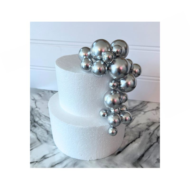 A two-tier white cake showcases a cascading cluster of Cake Sphere multi-sized balls in shiny Silver on one side. The cake is set against a backdrop with a white wainscot panel and rests on a marble surface.