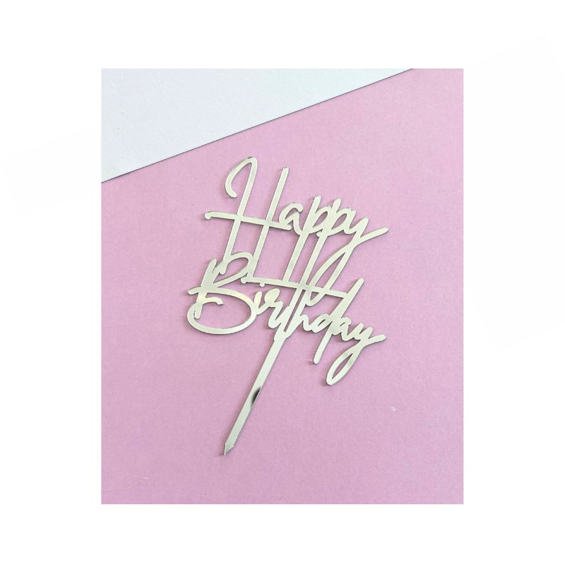 The "Silver Happy Birthday" cake topper features the words "Happy Birthday" written in an elegant, cursive font. Made of a metallic material, this topper is beautifully placed on a light pink surface.