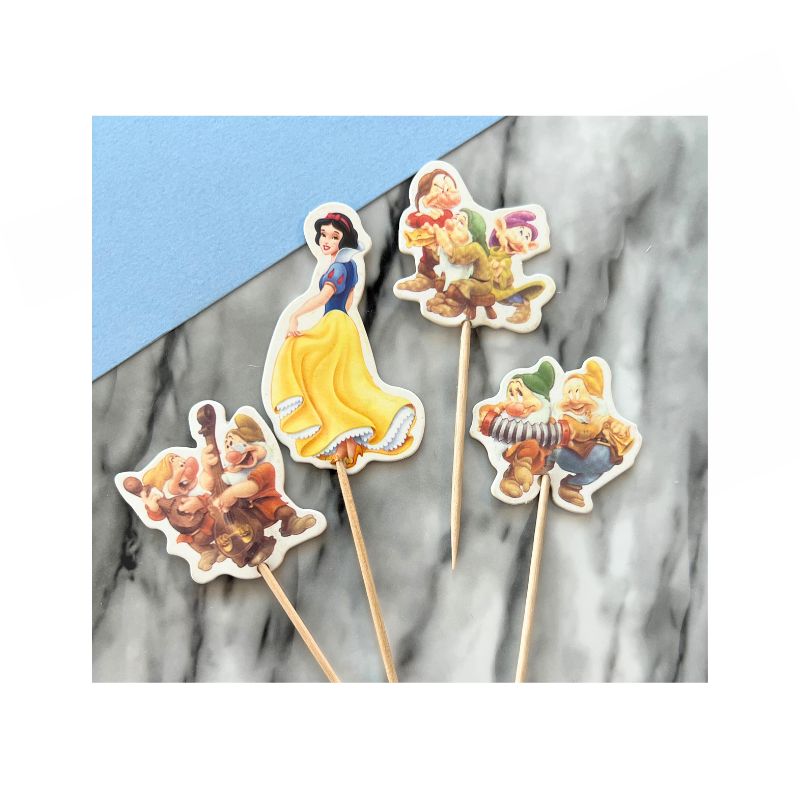 Four cupcake toppers feature characters from the well-known fairy tale "Snow White and the 7 Dwarfs": one shows Snow White in a yellow dress, while the others depict various characters in colorful outfits. The toppers have wooden picks and are displayed on a marble and blue surface.