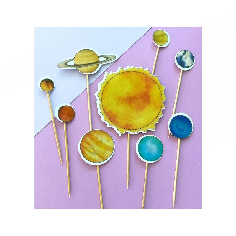 Introducing the Solar System Cupcake Toppers: a vibrant set of paper decorations featuring the planets of our solar system and the sun, each elegantly mounted on a toothpick. Displayed against a light pink and lavender dual-tone background, these toppers include Saturn, the sun, and various colorful planets, perfect for adding an astronomical touch to your treats.