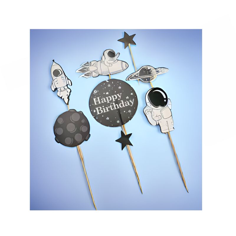 Space cake toppers on a blue background, showcasing astronaut, rocket, and planet designs. One topper displays a "Happy Birthday" message set against a round, starry backdrop. Each piece is attached to a wooden skewer and designed in black, white, and gray tones.