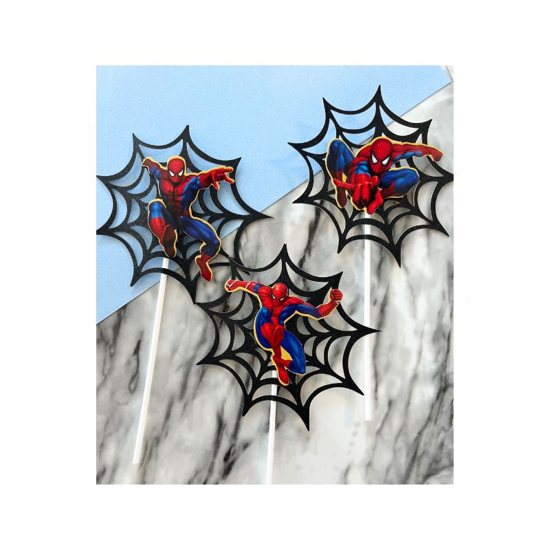 Three Spiderman cupcake toppers featuring the superhero in his iconic red and blue costume striking various action poses. Each topper has a black spider web background with a white stick attached, set against a light blue and gray marble-patterned surface.