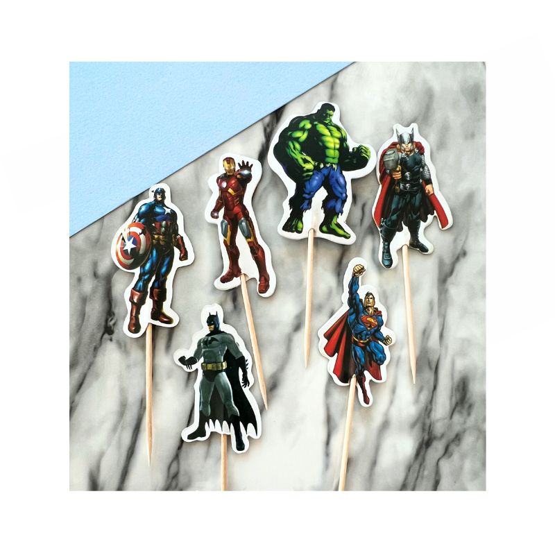 The Super Hero Cupcake Toppers set includes six characters: Captain America, Iron Man, Hulk, Thor, Batman, and Superman. Each topper is placed on a toothpick and arranged on a marble surface with a blue background in the corner.