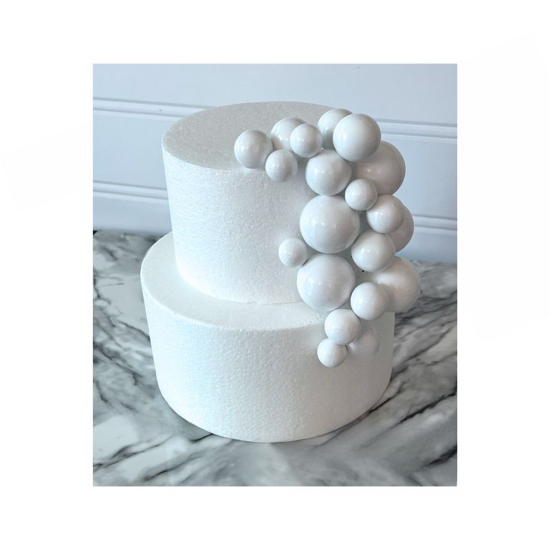 A two-tier white cake showcases a cascading cluster of Cake Sphere multi-sized balls in Whiteon one side. The cake is set against a backdrop with a white wainscot panel and rests on a marble surface.