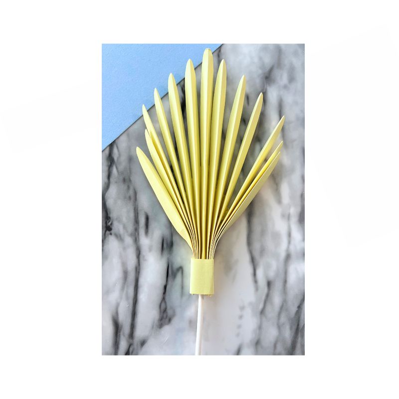 A Cake Paper Palm, fully opened with neat, symmetrical accordion folds, rests on a marble surface. The light yellow, folded paper fan with a handle partially overlaps a corner of pastel blue paper.