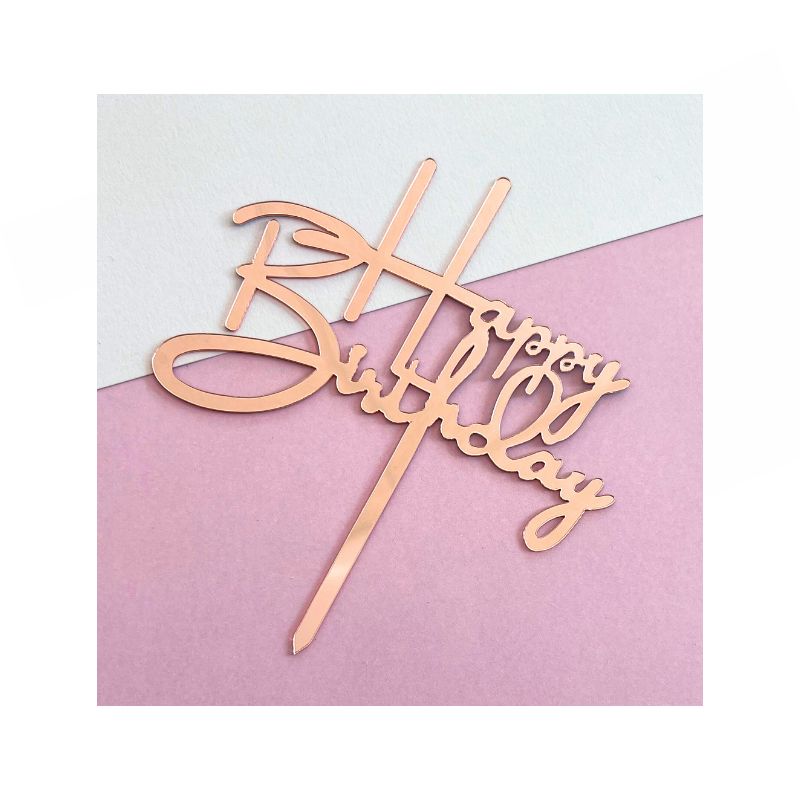 A Rose Gold Cake topper with elegant cursive text is placed on a two-toned background of light gray and pink. The topper's design includes a stick for easy insertion into a cake.