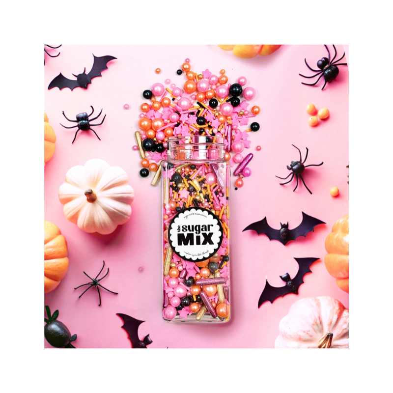 A rectangular jar labeled "Sassy but cute sprinkle mix" lies open on a pink surface, spilling Halloween-themed sprinkles. Surrounding it are plastic black bats and spiders, small pumpkins, and assorted pink, black, and orange candy beads.