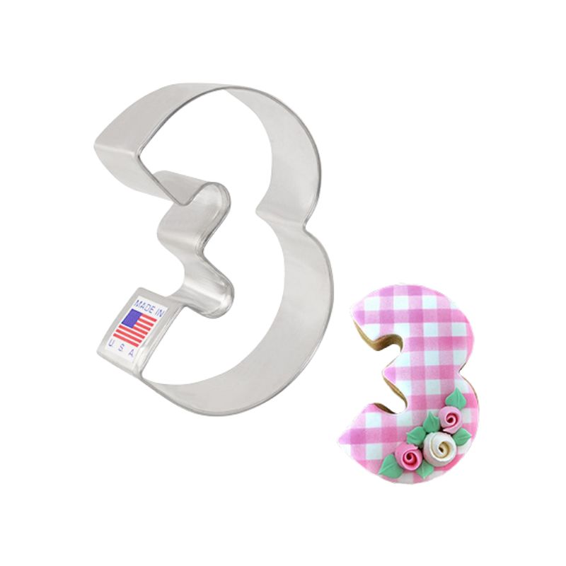 Metal cookie cutter in the shape of a three and made in the usa with flag sticker beside is a decorated cookie with pink gingham design and floral