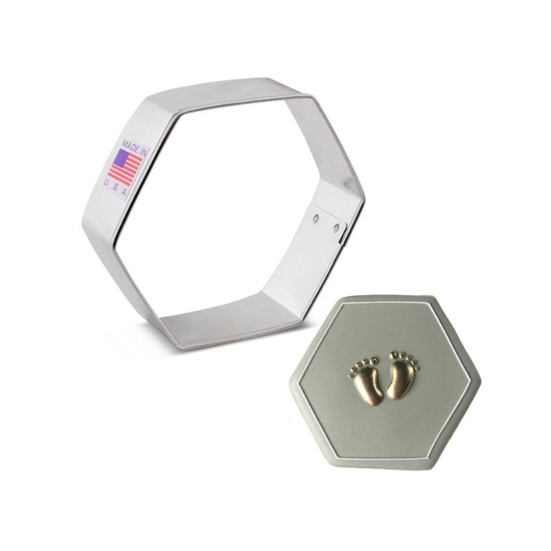 Stainless steel hexagon cookie cutter and beside is hexagon sugar cookie with gold feet on a taupe background