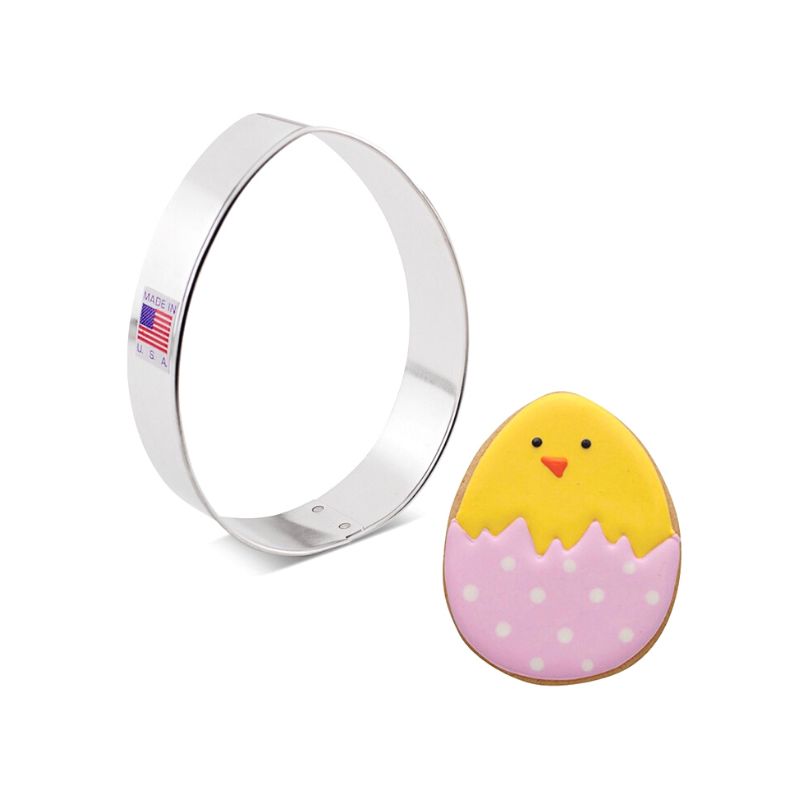 Stainless steel cookie cutter in the shape of an egg with a flag sticker on side beside a sugar cookie with pink polka dot shell and yellow duck on top with orange beak