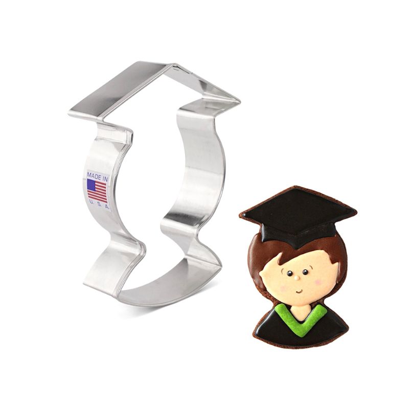 Stainless steel graduation person cookie cutter with flag sticker is next to decorated grad person with black grad hat and gown with green lapels and boy face