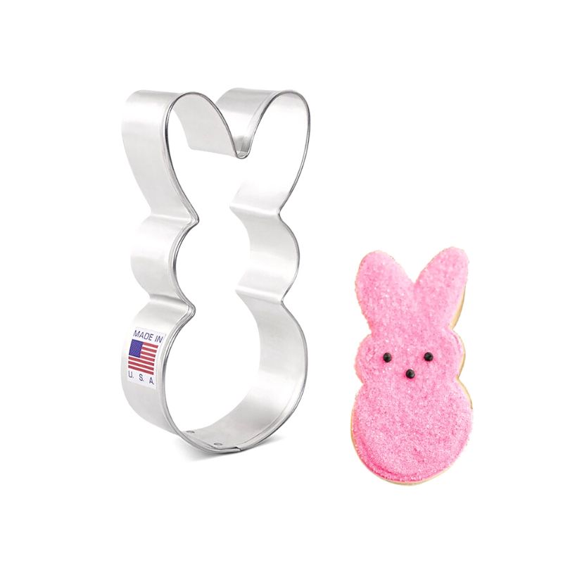 Stainless steel peeps bunny cookie cutter with made in USA and flag sticker on side next to pink decorated sugar cookie with black eyes and nose