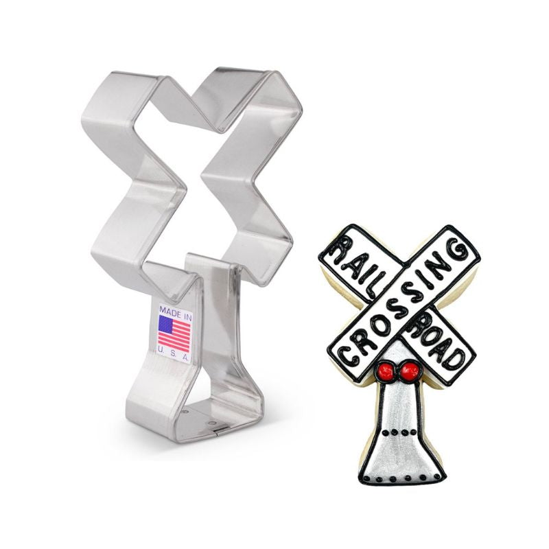 Metal railroad crossing cookie cutter with made in USA flag sticker on the side next to sugar cookie with railroad crossings in black icing and silver base with red  icing lights