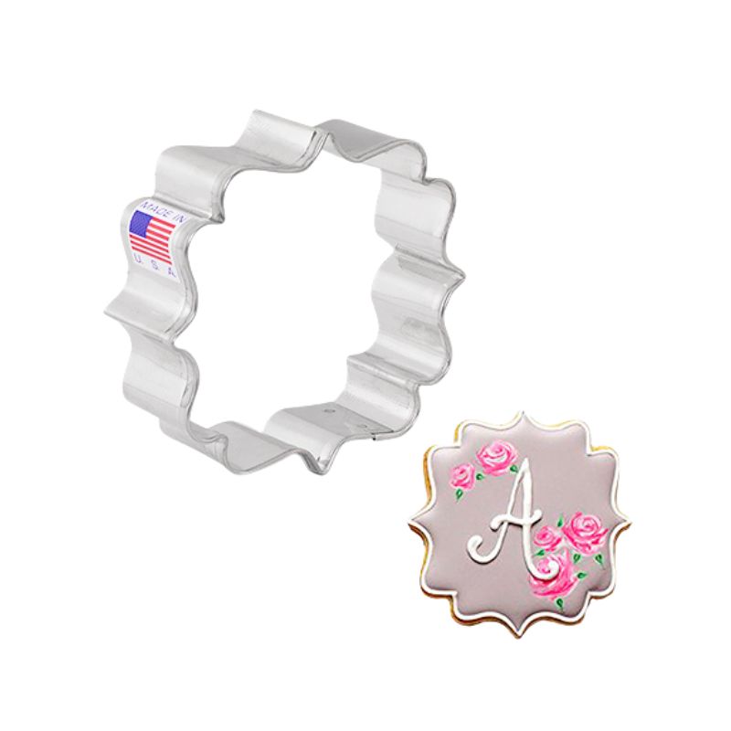 Metal cookie cutter square with scallop edges made in the USA next to a decorated plaque cookie with taupe royal icing and pink flower